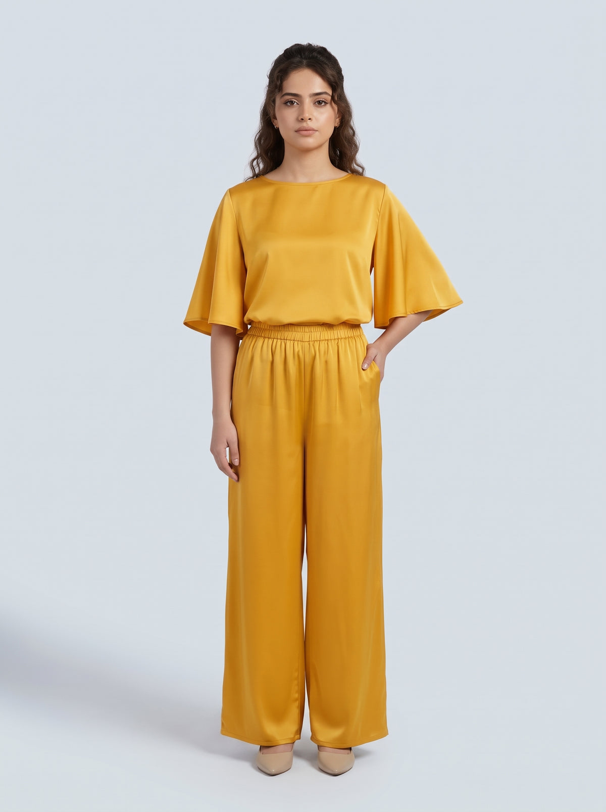 A woman modeling a matching two-piece outfit consisting of a mustard yellow short-sleeve top with flared sleeves and matching wide-leg trousers, set against a solid light gray background.