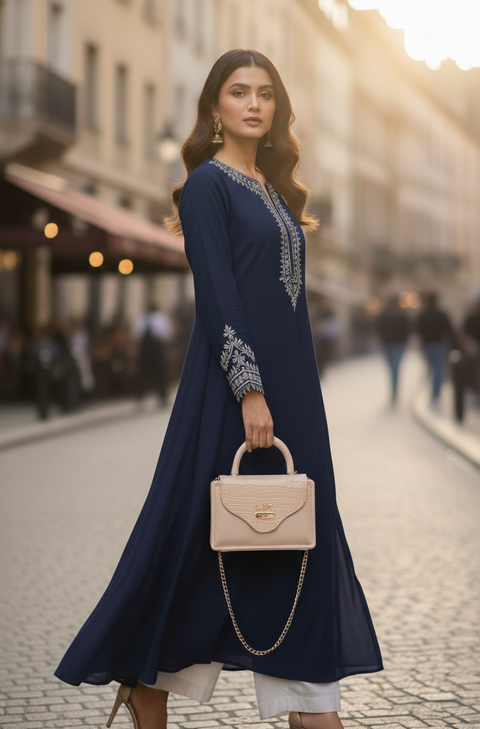 Woman in a long navy dress with white embroidery holding a beige handbag on a city street.