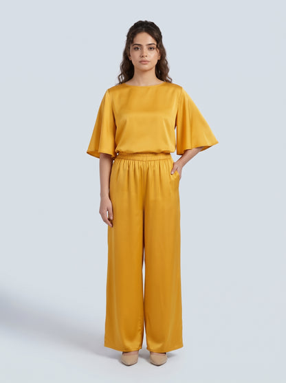 A woman modeling a matching two-piece outfit consisting of a mustard yellow short-sleeve top with flared sleeves and matching wide-leg trousers, set against a solid light gray background.