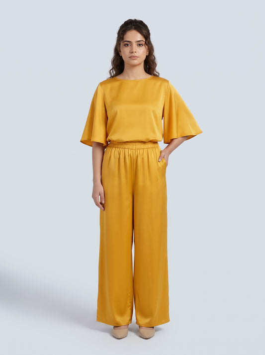 A woman modeling a matching two-piece outfit consisting of a mustard yellow short-sleeve top with flared sleeves and matching wide-leg trousers, set against a solid light gray background.